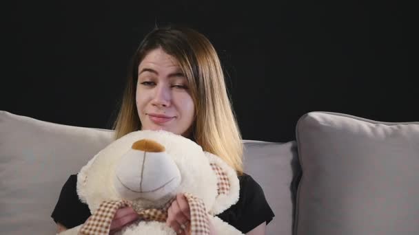 Pretty smiling woman sitting on the couch hugging teddy bear - Imágenes, Vídeo