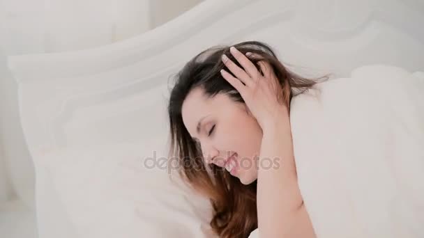 Young brunette woman lying on the bed and playing with hair in light apartment. Girl looks at camera and cute smile. - Záběry, video