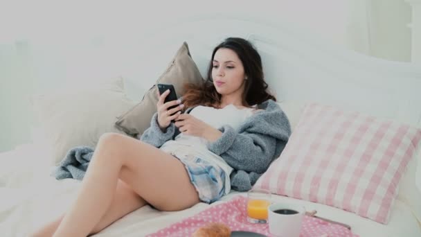 Young woman using smarphone during breakfast lying on bed at home. Attractive girl surfs the Internet on mobile phone. - Video