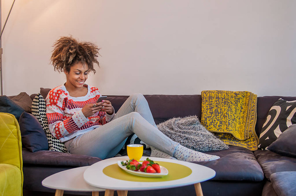 Happy woman at home reading a text message in her bright livingroom. Relaxed young woman text messaging on sofa - Fotografie, Obrázek