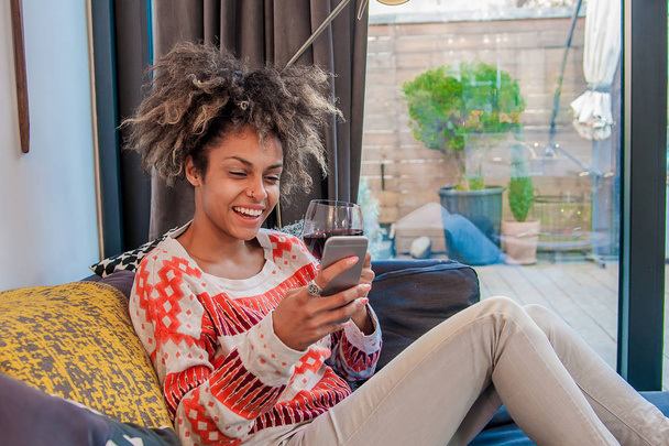 Menina jovem hipster usando o dispositivo de smartphone moderno enquanto se senta em casa e se comunica com amigos em redes sociais, freelancer do sexo feminino trabalhando em casa, felicidade e estilo de vida de prazer
 - Foto, Imagem