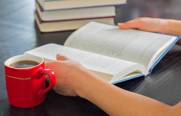 Mulher mãos segurando livro aberto com pilha de livros fechados e xícara de café em casa
.. - Foto, Imagem