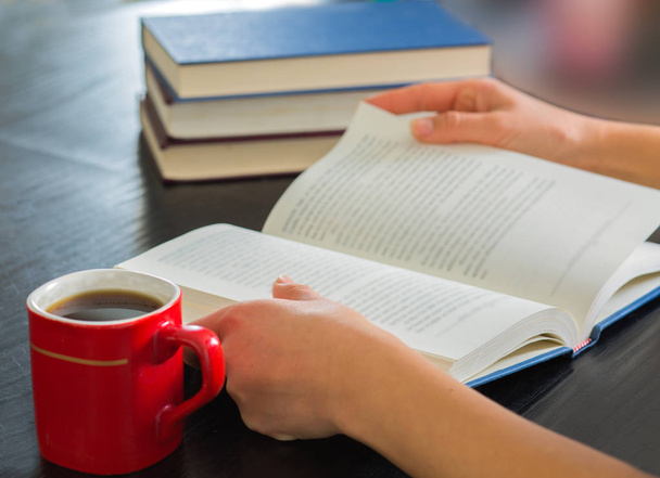 Manos de mujer sosteniendo libro abierto con pila de libros cerrados y taza de café en casa
.. - Foto, imagen