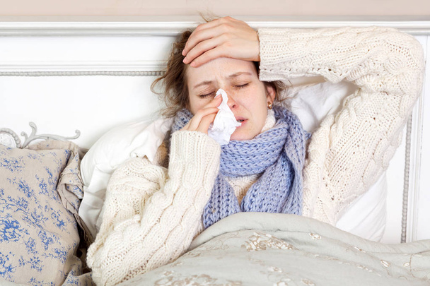 Flu. Closeup image of frustrated sick woman lying in bed in thick blue scarf holding tissue by her nose and touching her head with closed eyes. - Photo, image
