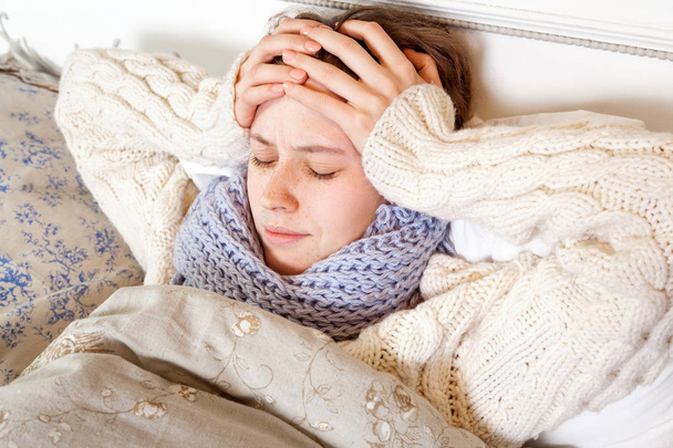Flu or cold. Closeup top view image of frustrated young woman with blue scarf and suffering from terrible headache while lying in bed. - Zdjęcie, obraz