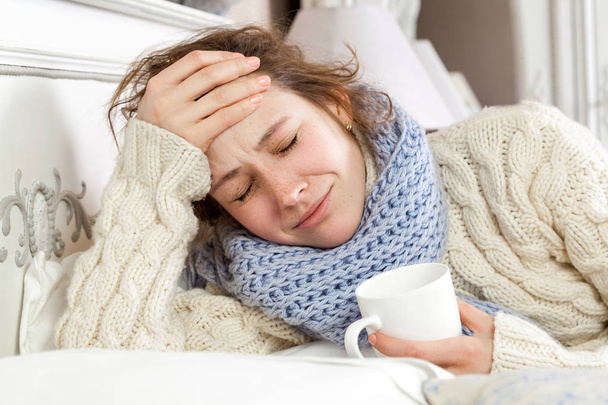Sick woman with cup of tea. Closeup image of young frustrated sick woman in knitted blue scarf holding a cup of tea while lying in bed. hand on head, sad and closed eyes.  - Foto, Bild