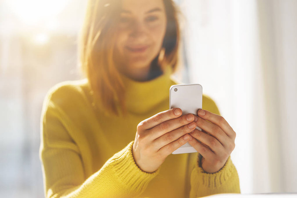 Imagen de cerca de la joven hipster sentada en el interior de su casa y utilizando un dispositivo de teléfono inteligente moderno
 - Foto, imagen