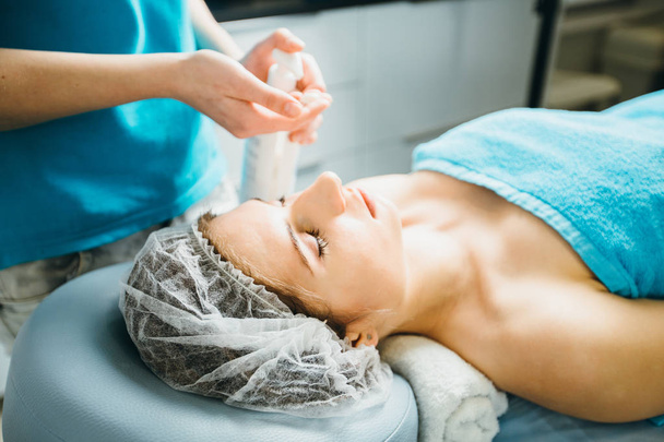 Les mains de l'esthéticienne pressent la crème pour le corps debout près d'une jeune femme allongée sur une table de massage dans un salon de beauté sain intérieur
. - Photo, image