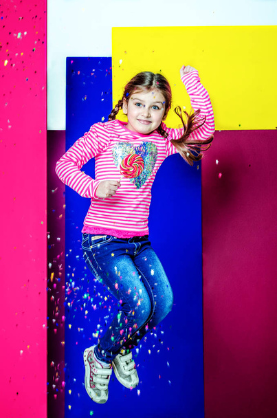 A girl in a pink blouse and blue jeans holds a candy in her hands and bounces. Confetti is spreading over the girl. - Photo, Image