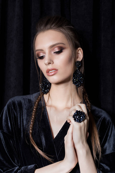 beautiful sexy young woman with dark hair braided with bright makeup and fashion bizhuterieyker earrings and ring, fashion jewelry shooting on a black background in the studio, hard light - Foto, immagini
