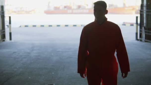 Young worker in orange uniform walking through the harbour facilities by the sea during his break. Leisure time - Materiał filmowy, wideo