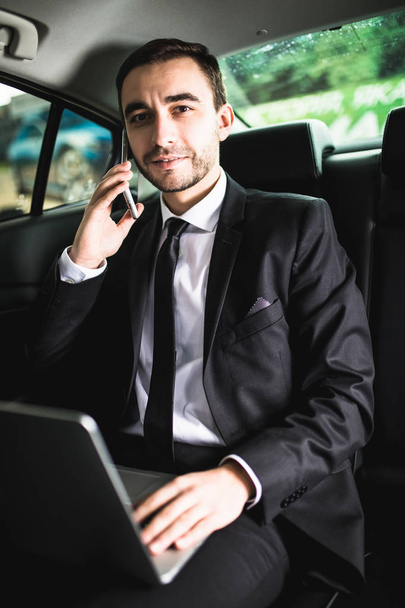 Handsome young businessman working on his laptop and talking on the phone while sitting in the car - Fotoğraf, Görsel