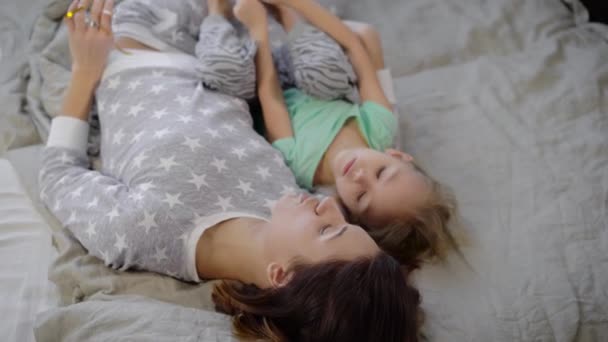 Mother with the daughter in a bedroom. The girl with the child lie on a bed and talk. The baby in a t-shirt of mint color and gray panties. The woman in a pajamas with stars. Young family. - Video, Çekim