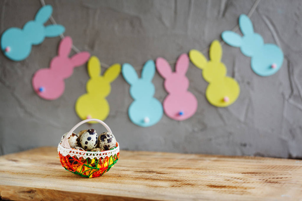 Basket with quail eggs and colorful bunnies garland on wooden background - Valokuva, kuva