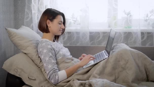 Portrait of beautiful brunette woman lying in bed with laptop at home in the bedroom. Young girl with short haircut is working as a freelancer spending her business day in the room. - Filmati, video