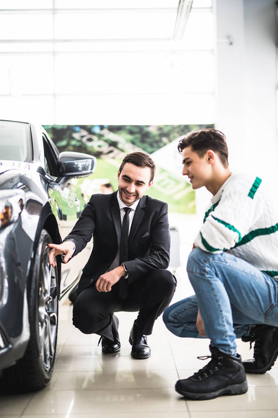 junger Autoverkäufer zeigt dem Kunden die Vorteile des Autos und der Reifen. - Foto, Bild