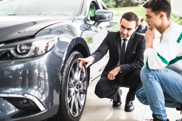 Молодой продавец автомобилей, показывающий преимущества автомобиля перед клиентом и шины
. - Фото, изображение