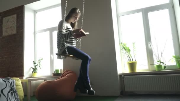 Teenager girl sitting on swind indoors and reading a book - Materiał filmowy, wideo
