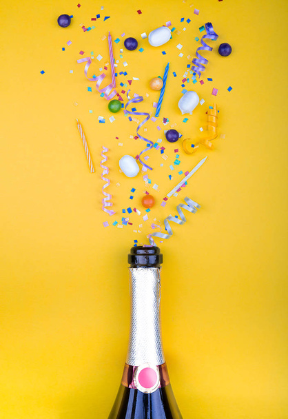 Colorful party attributes fly out from botle of champagne wine. on a yellow background. Flat lie. Celebrate concept. High resolution photo. - Fotoğraf, Görsel