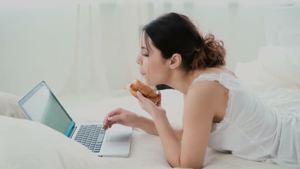 Beautiful girl lying on the bed in morning, using laptop. Young woman eating croissant, typing, browsing the Internet. - Imágenes, Vídeo