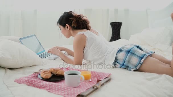 Young woman using laptop during breakfast lying on white bed at home. Brunette girl typing on pc and eating croissant. - Video