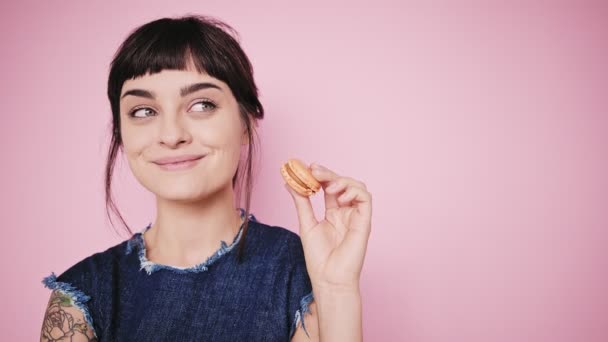 Atrakcyjna brunetka zjada macarons na białym tle na jasnym tle - Materiał filmowy, wideo
