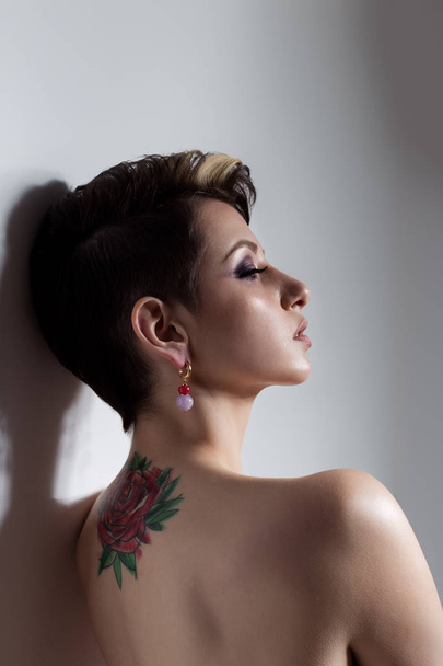 Beautiful young sexy girl with short hair with tattoo on his back is against the wall with bare shoulders sad - Photo, Image