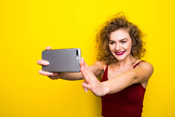 Young beautifulgirl with an curly hairstyle. Laughing girl take selfie from phone on Yellow background. - Foto, Imagem