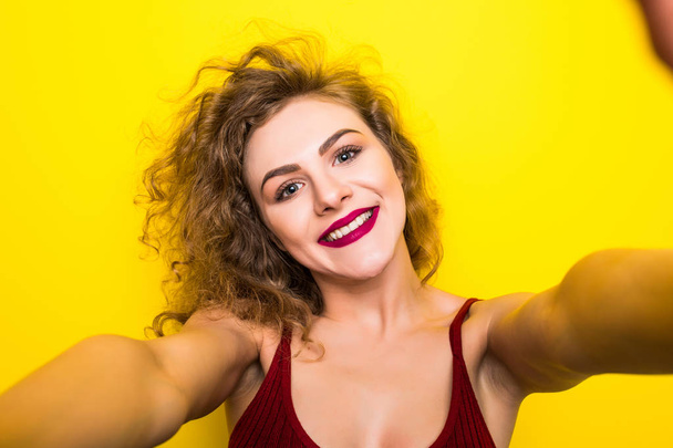 Young beautifulgirl with an curly hairstyle. Laughing girl take selfie from phone on Yellow background. - Photo, image