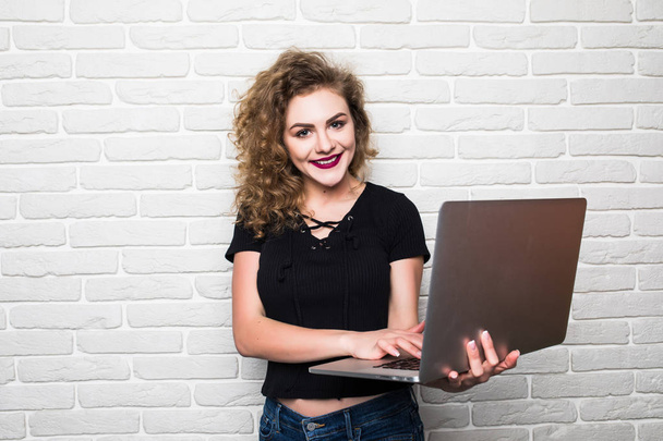 Bella giovane donna in abbigliamento casual sta utilizzando un computer portatile e sorridente, in piedi contro muro di mattoni bianchi
 - Foto, immagini