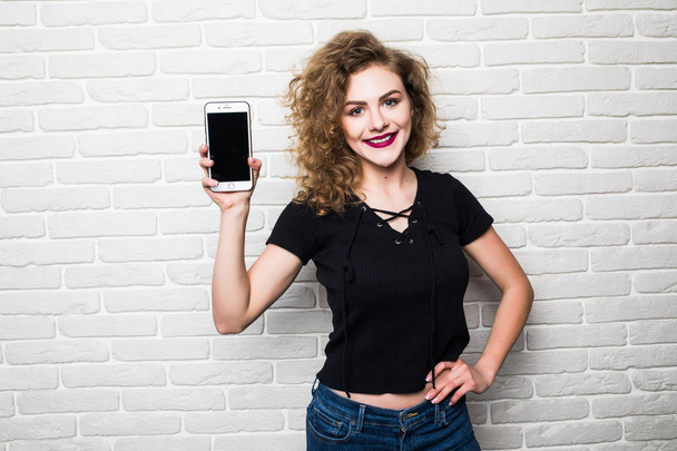 Mooie jonge vrouw met kopie ruimte slimme telefoon, tegen witte bakstenen muur - Foto, afbeelding
