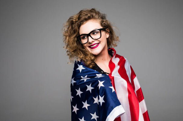 Cheerful young woman in casual clothes and glasses is covered in the American flag and smiling on gray background - Foto, imagen