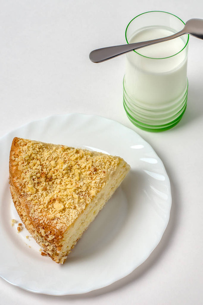 Кусок печенья нежный торт на белой тарелке на столе на светлом фоне, избирательный фокус. Сверху. Слоистый торт покрыт крошками, рядом с десертом металлическая винтажная ложка и стакан белого молока
. - Фото, изображение