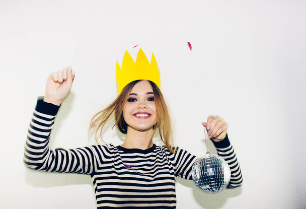 Birthday party, new year carnival. Young smiling woman on white background celebrating brightful event, wears stripped dress and yellow crown. Sparkling confetti, having fun, dancing, laugh, smile. - Foto, imagen