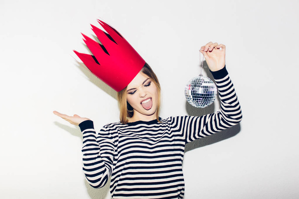 Young smiling woman celebrating party, wearing stripped dress and red paper crown, happy dynamic carnival disco ball party, excited, having fun, smiling, laugh. White background - Photo, image