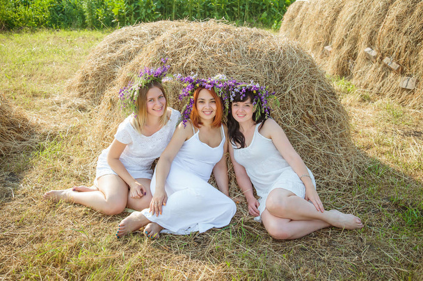Three young beautiful girls in white dresses and wreaths of wild flowers sitting near haystack. Summer in the village - Photo, Image
