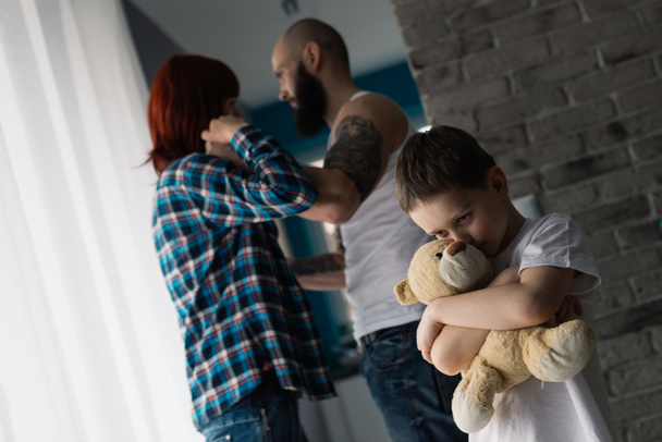 Грустный ребенок обнимает своего плюшевого мишку во время ссоры родителей
. - Фото, изображение
