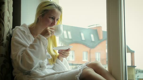junges Mädchen im Bademantel trinkt Kaffee zu Hause am Fenster - Filmmaterial, Video