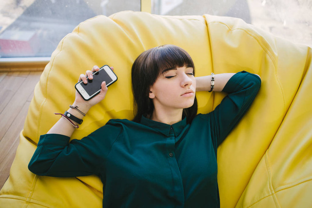 Giovane bella donna che dorme durante la pausa in una comoda sedia e in possesso di un telefono. Ritratto di un ufficio moderno
 - Foto, immagini