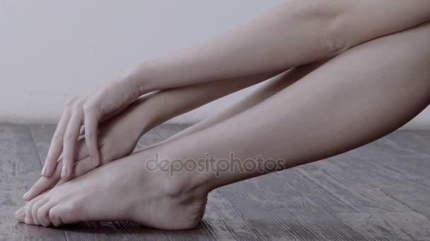 Donna delicatamente toccando le gambe seduto sul pavimento in legno con parete bianca su sfondo
. - Filmati, video