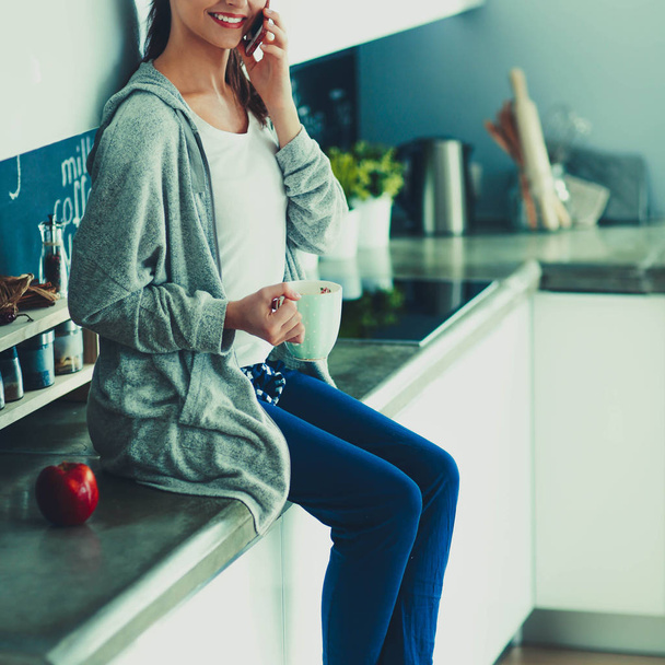 Woman using mobile phone sitting in modern kitchen - Photo, Image