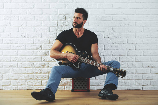 Handsome european man in casual clothing playing the guitar while standing in interior with white brick wall and wooden floor. Art concept - Fotó, kép