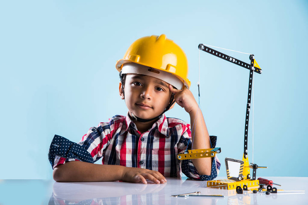 cute indian baby boy playing with toy crane wearing yellow construction hat or hard hat, childhood and education concept - Photo, Image