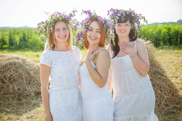 Три юные красивые девушки в белых платьях и венки из диких цветов, стоящие возле стога сена и смеющиеся. Лето в деревне
 - Фото, изображение