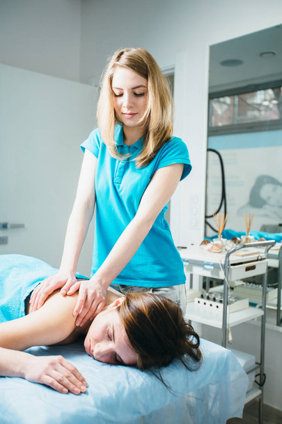 Mujer joven acostada vestida con toalla turquesa mientras masajea a su terapeuta de masajes en los hombros
 - Foto, Imagen