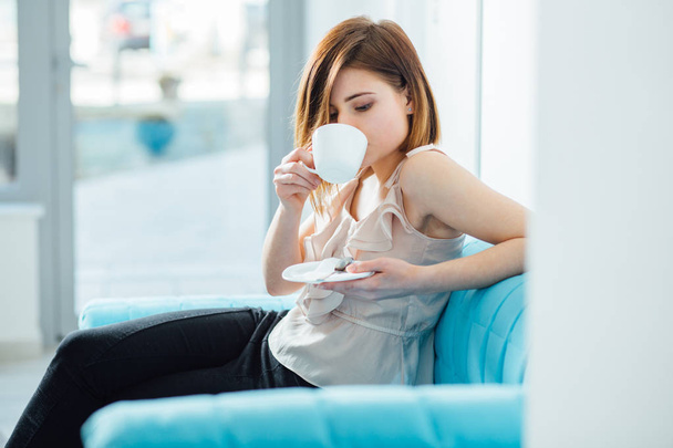 Portrait of female visitor waiting for a doctor in hall of clinic while sitting in turquoise sofa and drinking coffee or tea from white cup on bright lighting background. - Fotoğraf, Görsel