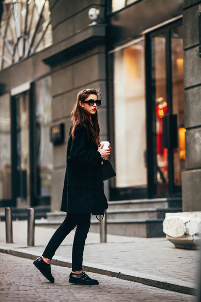 young stylish girl, passing by the Windows.Wearing fashionable glasses and a black coat.Keeps coffee.Abstract looking to the side. - Foto, Bild