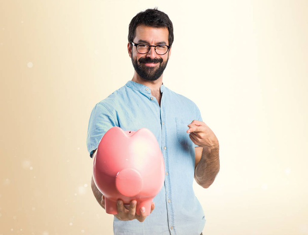 Handsome man with blue glasses holding a piggybank on ocher back - Photo, Image
