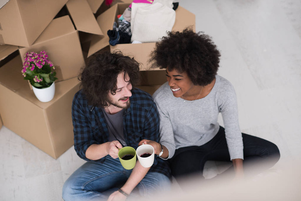 Relaxing in new house. Cheerful young multiethnic couple sitting on the floor and drinking coffee while cardboard boxes laying all around them - Foto, Imagen