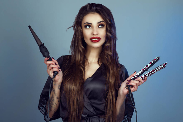 Beauty portrait of young gorgeous smiling model with perfect teeth and long hair wearing black silk peignoir holding curling wand and hair straightener - Photo, Image